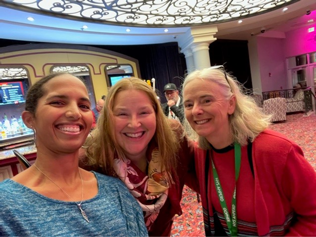 Three smiling people posing indoors under a decorative ceiling, with a bar and neon lights in the background.