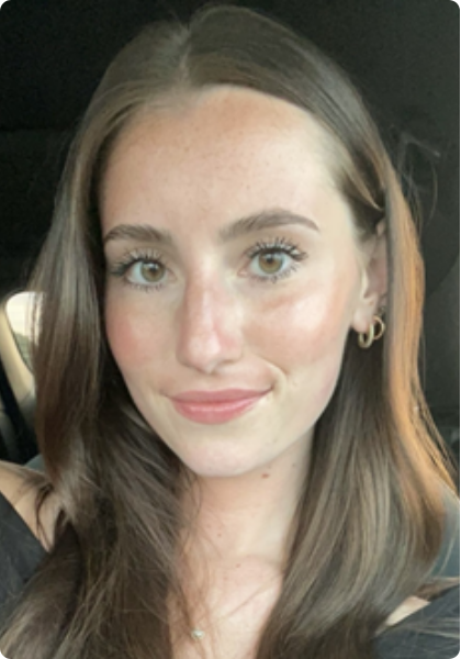 Close-up selfie of a smiling person with long brown hair, wearing a black top and gold hoop earrings in a car.