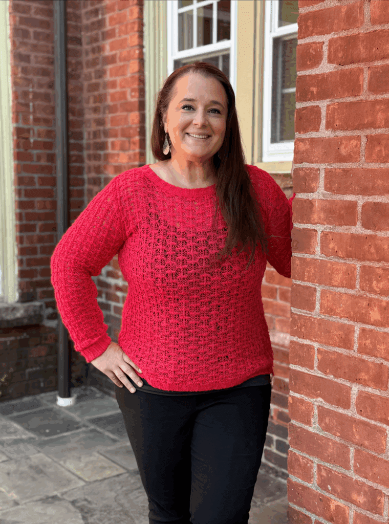 Woman in a bright pink sweater and black pants standing by a brick wall, smiling at the camera.