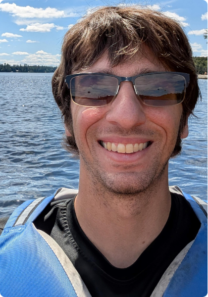 Smiling person in sunglasses and a blue life jacket on a boat with water and a cloudy sky behind them