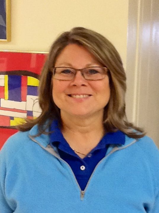 Smiling woman in glasses and blue sweater, standing indoors near a colorful abstract painting