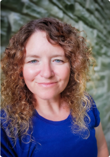 Smiling woman with curly hair in a blue shirt, standing outdoors against a blurred green background