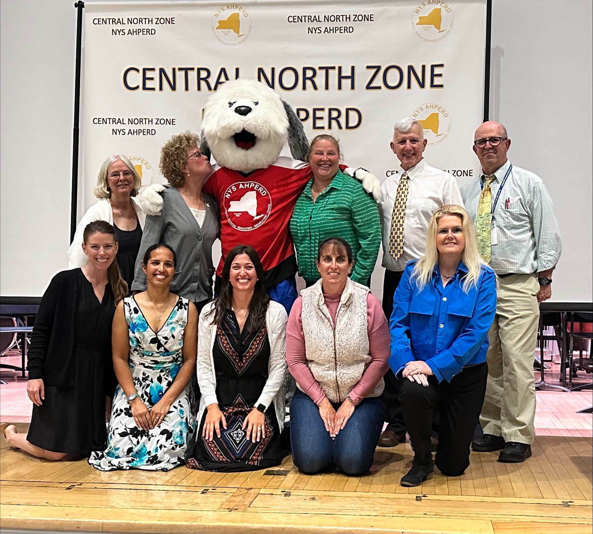 Group posing with a bear mascot in front of a “Central North Zone” banner on a stage.
