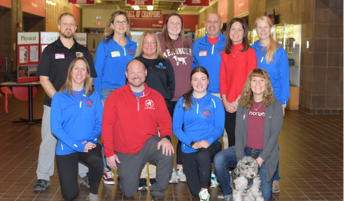 Group of volunteers in colorful sweatshirts posing indoors in a lobby with a small dog in front