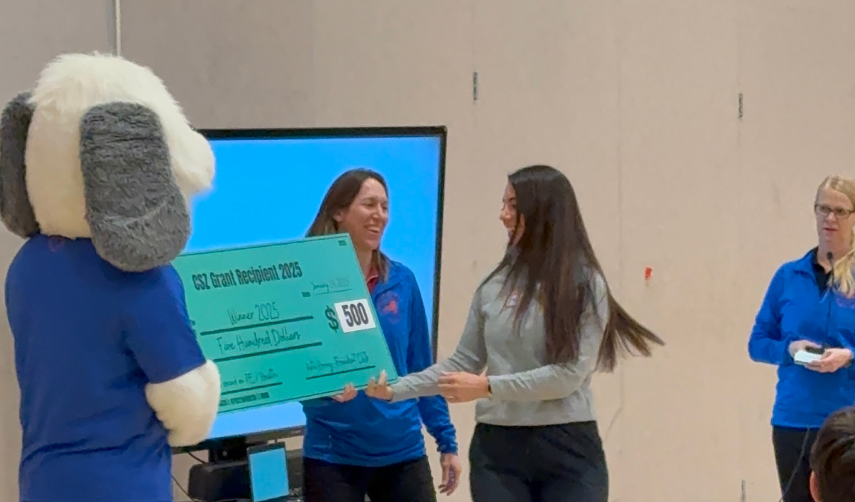 Mascot presenting a large check to a woman at an indoor event while another woman watches nearby