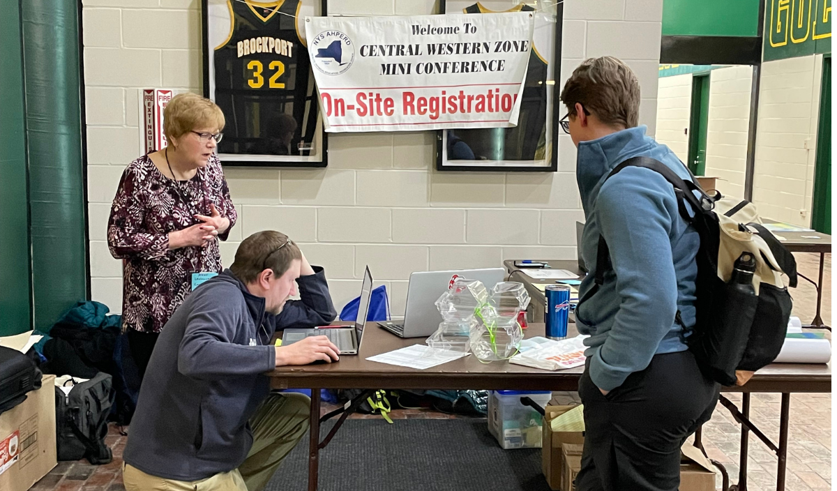 People at a table indoors under a “Pie Site Registration” sign, chatting and working with papers and drinks.