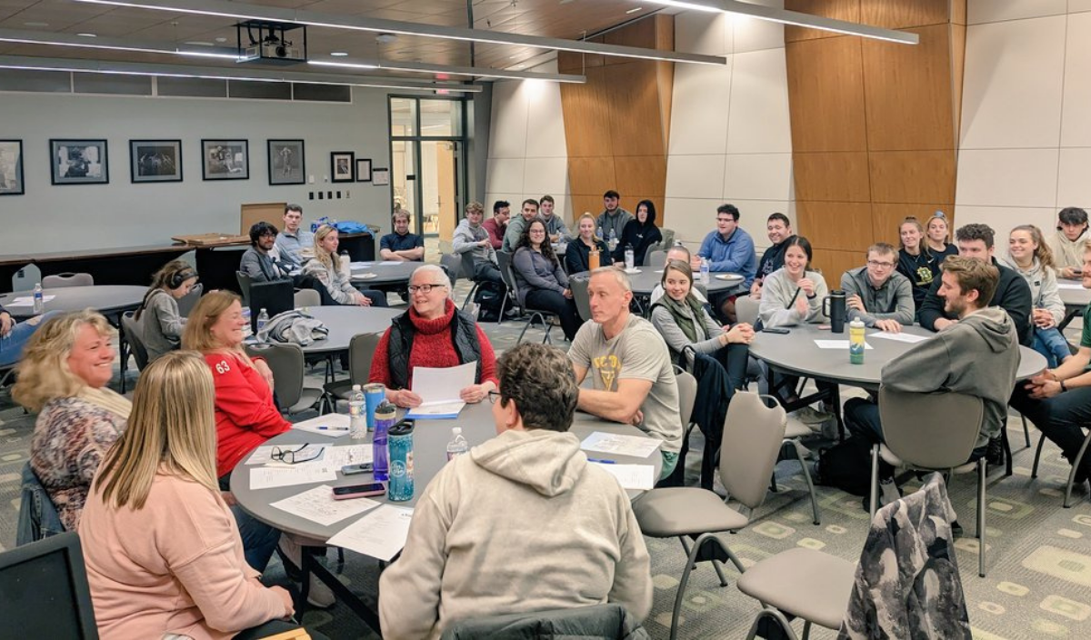 People seated at tables in a community meeting room, listening and talking during a group discussion.