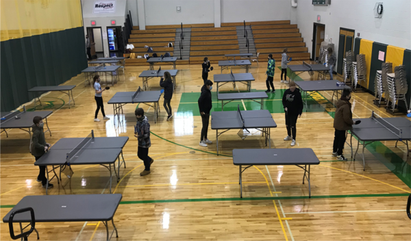 People playing table tennis in a gymnasium with multiple green and gray tables