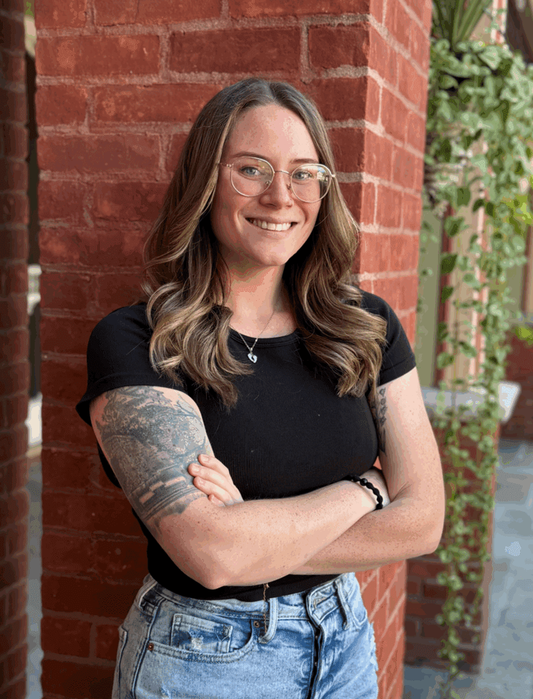 Smiling person with glasses and tattoos, arms crossed, standing by a red brick wall outdoors