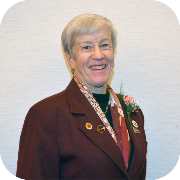 Smiling person in a burgundy blazer with medals and floral pin against a plain light background