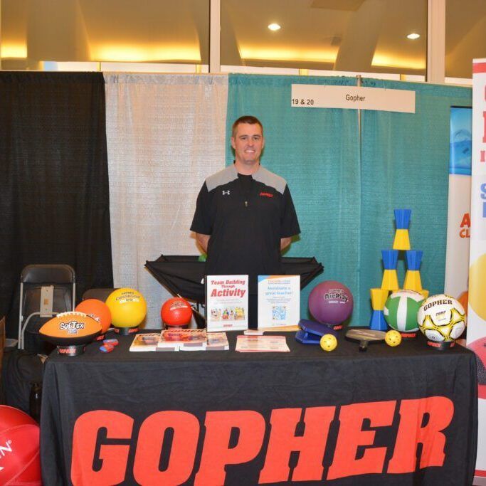 Man standing behind a Gopher Sports display table with colorful balls and training equipment at an indoor expo
