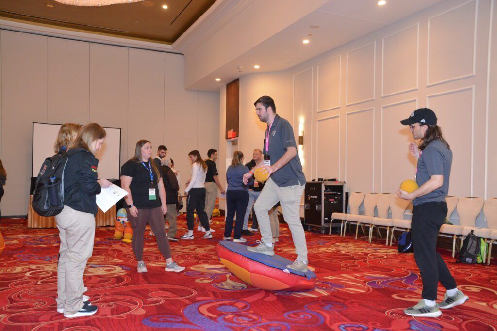People balancing on foam wedges in a carpeted conference room, with others watching nearby.