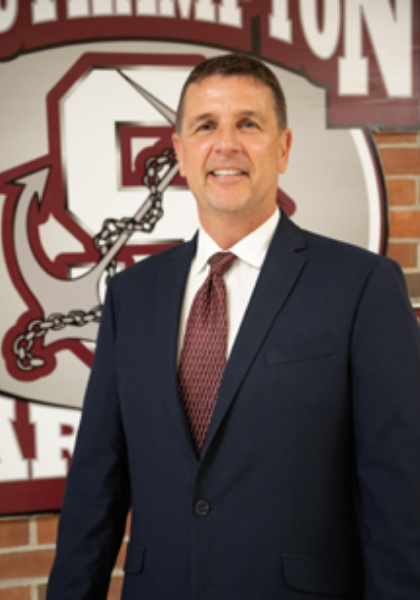 Man in a navy suit and red tie standing before a maroon-and-white team logo backdrop