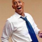 Smiling man in a white shirt and blue tie, leaning back with an open-mouthed laugh