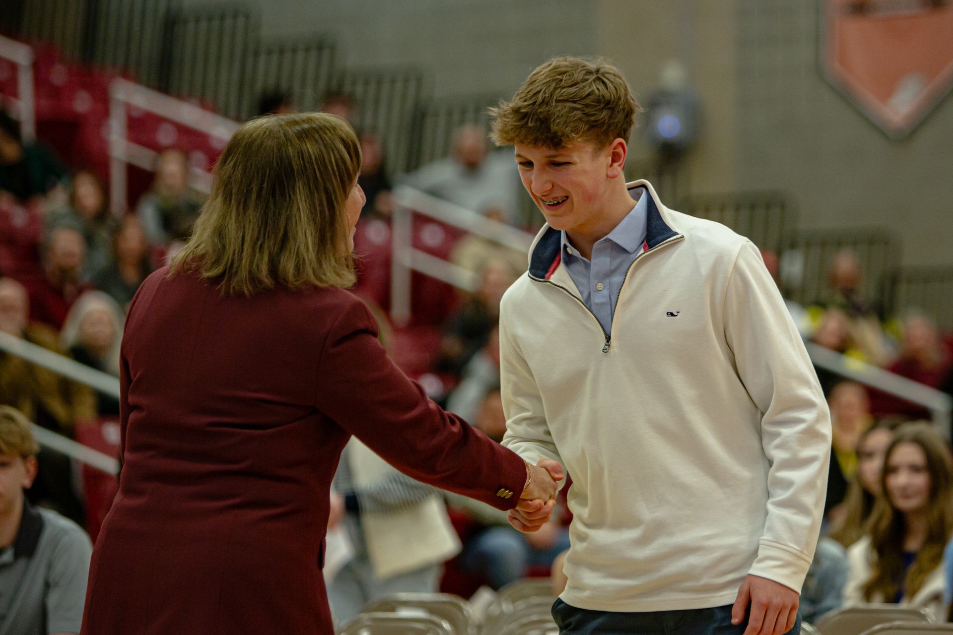 Two people shake hands in a gymnasium, with spectators seated in the background.