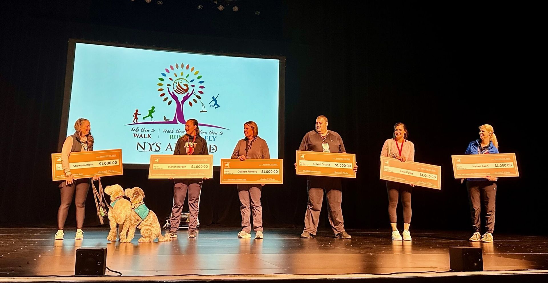 Six people on a stage holding cardboard signs, with two dogs in front of a colorful presentation screen.