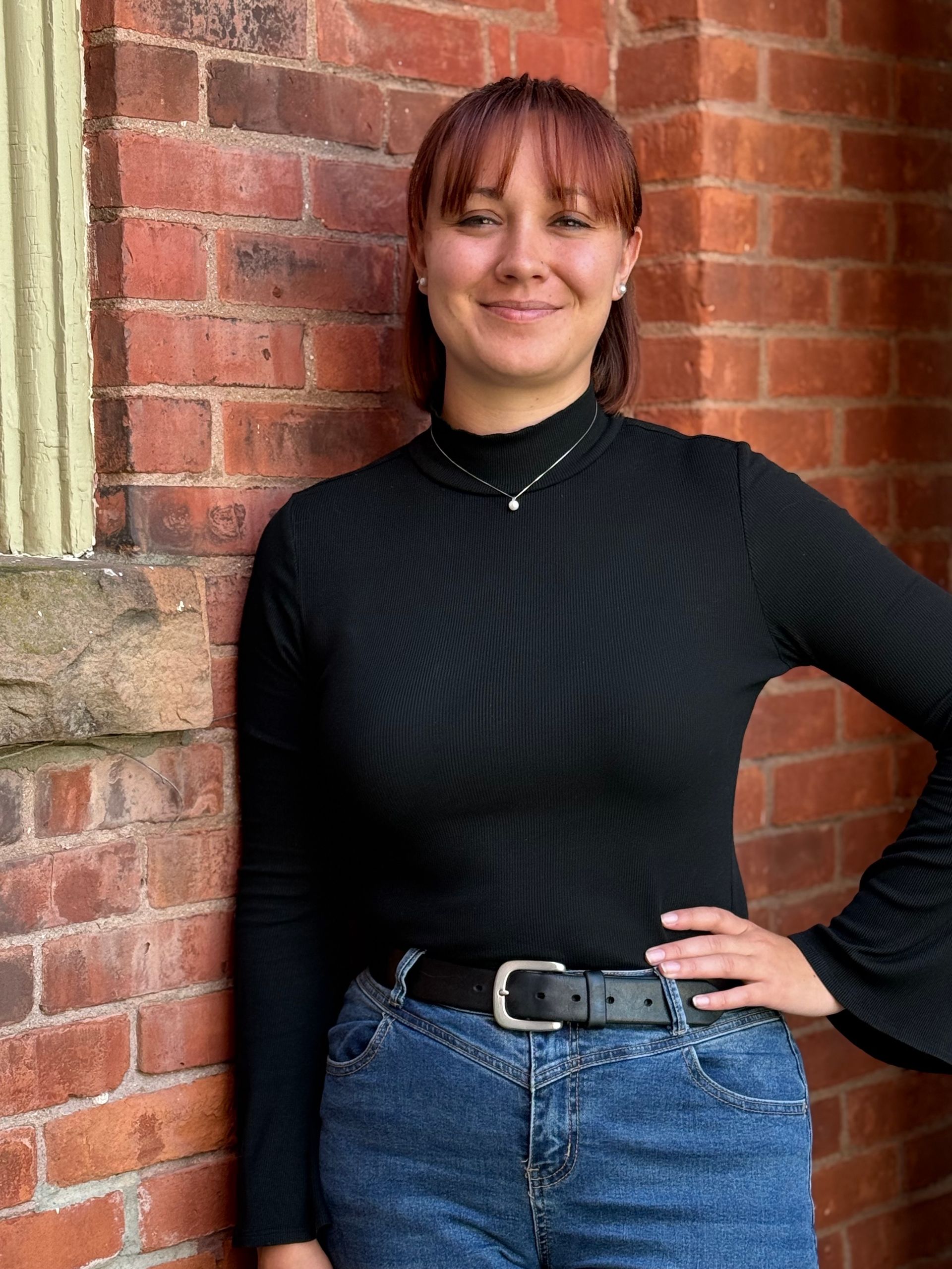 Woman leaning against a brick wall, wearing a black top and blue jeans, smiling at the camera.