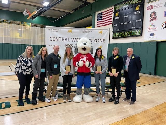 Group posing on a gym court with a mascot in front of a Central West Zone banner