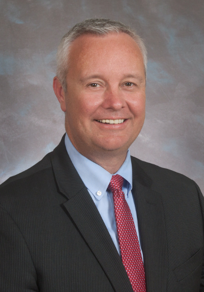 Portrait of a smiling man in a dark suit, light blue shirt, and red tie against a gray studio background