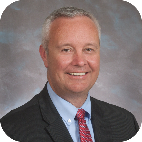 Formal studio headshot of a smiling man in a dark suit, light blue shirt, and pink tie