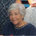 Smiling woman with short white hair in a blue sweater, seated indoors against patterned fabric.