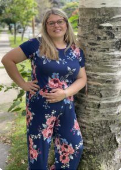 Woman in a navy floral dress standing by a birch tree in a park, smiling with one hand on her hip.