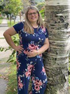 Woman in a navy floral jumpsuit standing beside a birch tree on a sidewalk, smiling with hand on hip.