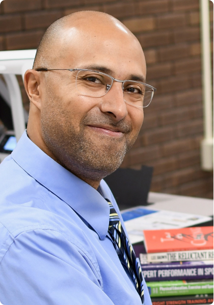 Smiling man in glasses and blue shirt seated indoors at a desk with books and a brick wall behind him