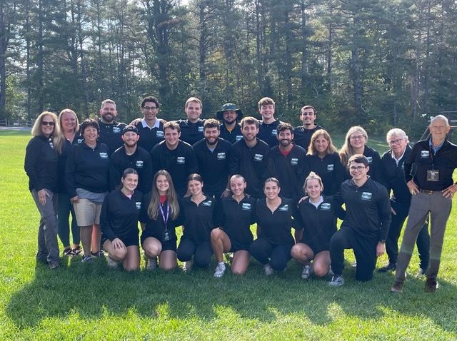 Group of people posing outdoors on a grassy field, wearing dark sports uniforms with trees in the background