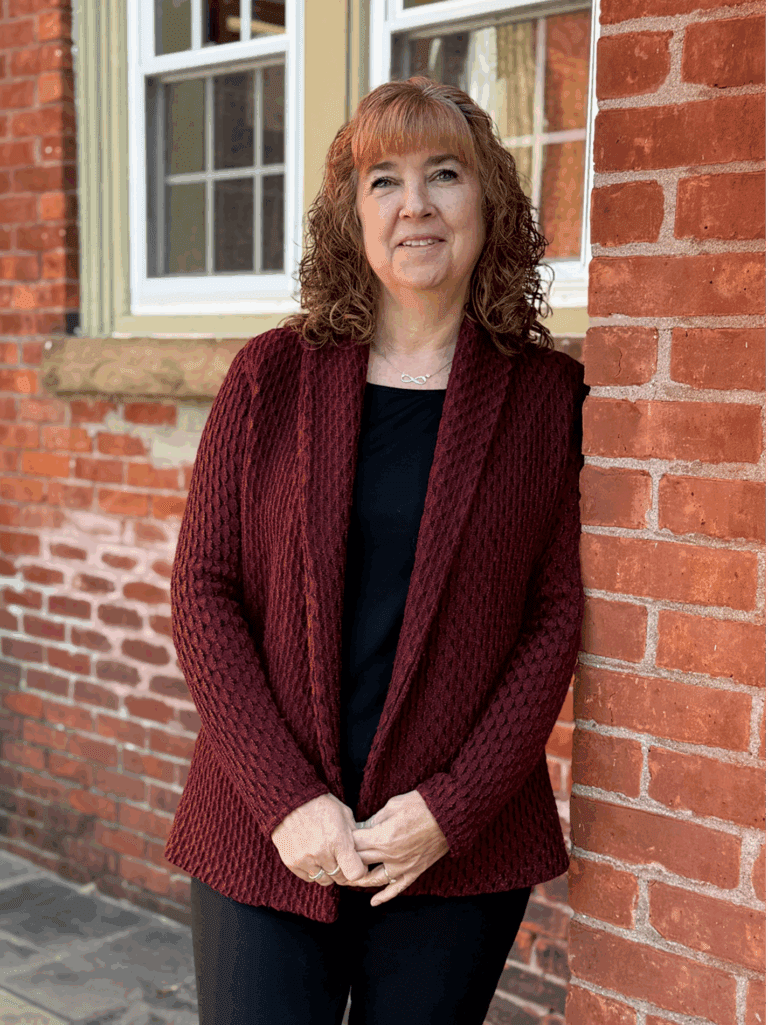 Woman in a maroon shawl standing by a brick wall and window outdoors.