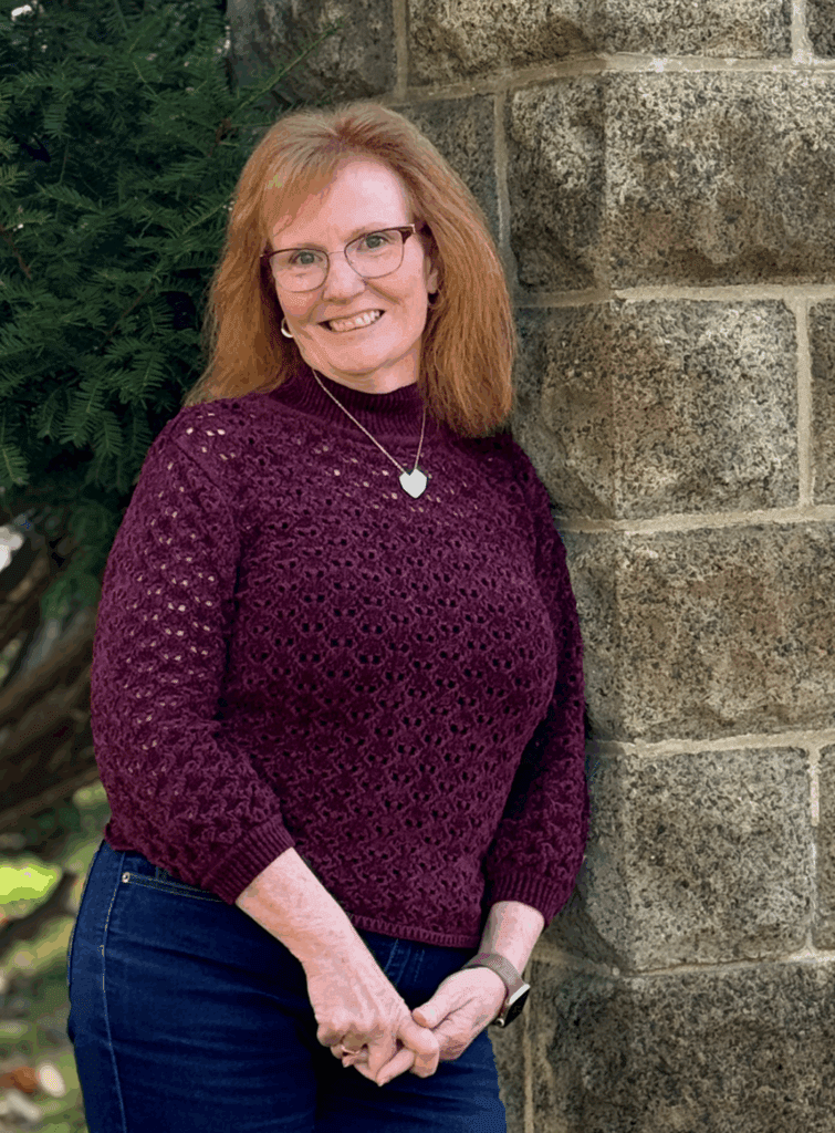 Woman in a burgundy sweater standing by a stone wall, smiling at the camera.