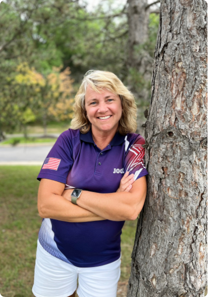 Woman in a purple polo shirt leans against a tree in a park, smiling at the camera.