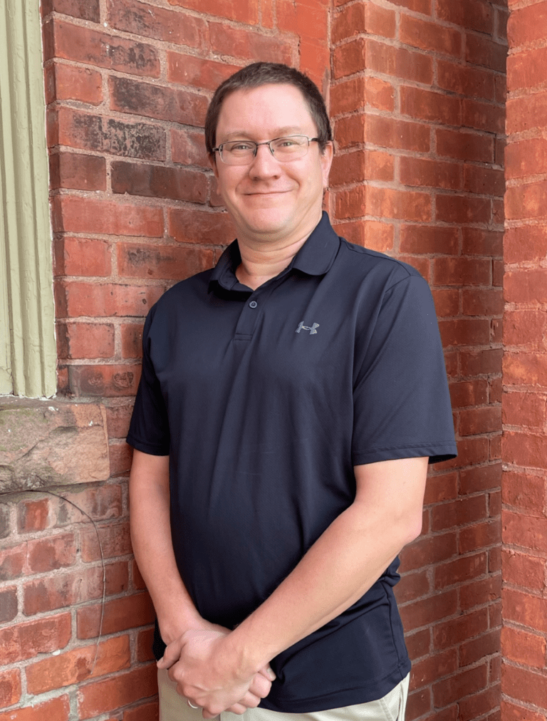 Person in a dark polo shirt standing against a brick wall, smiling with hands clasped.