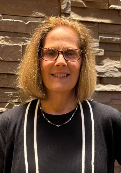 Smiling person wearing glasses and a black top with white trim, standing before a stone wall.