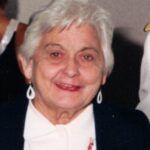 Smiling elderly woman with short white hair, wearing a white top and black jacket