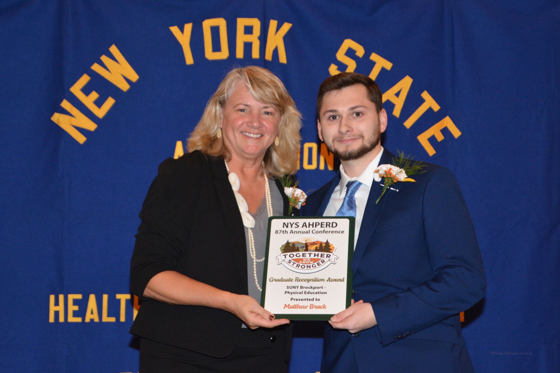 Two people hold a framed award in front of a New York State Health backdrop.