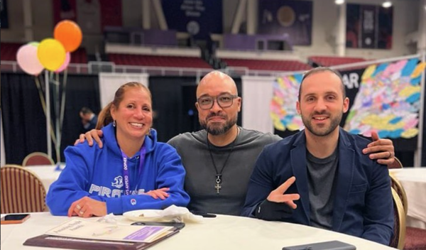 Three people smiling at a table indoors, with balloons and colorful artwork in the background.