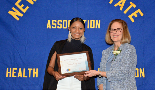 Two women hold a framed certificate in front of a blue association banner.