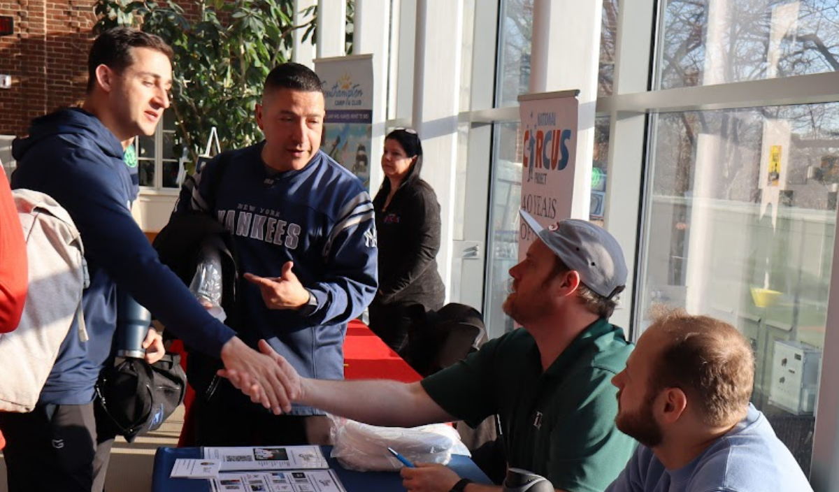 People talking and shaking hands at a table outside a storefront, with papers and signs visible.