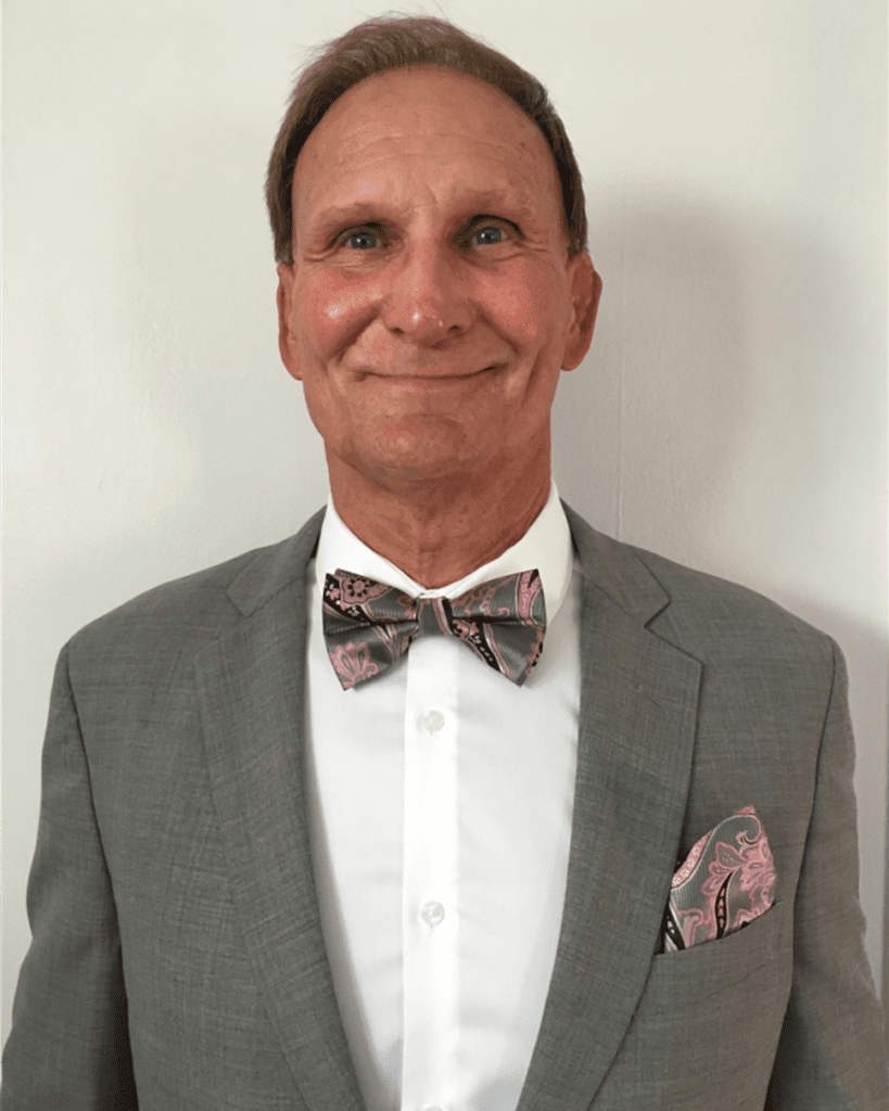 Man in a gray suit with a floral bow tie and pocket square, standing against a plain light background.