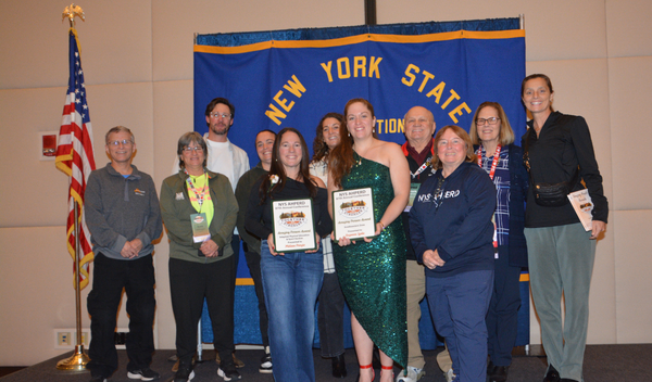 Group of people holding certificates in front of a New York State banner at an indoor event