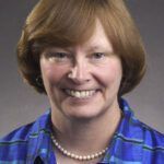 Smiling woman with short light-brown hair, wearing a blue jacket and pearl necklace against a gray background