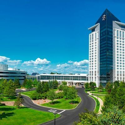Modern office campus with a tall glass tower, landscaped lawns, and winding roads under a bright blue sky