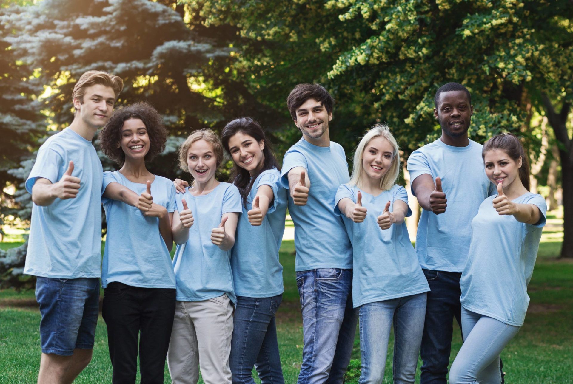 Group of people in light blue shirts standing outdoors on grass, smiling and holding drinks toward the camera