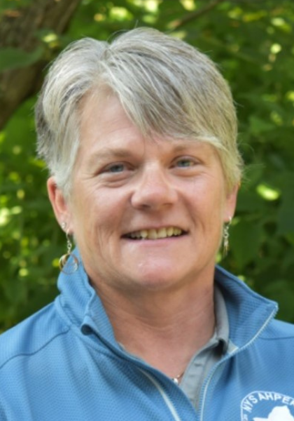Smiling person with short gray hair in a blue jacket, outdoors with green foliage background