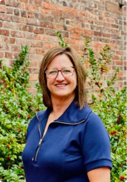 Woman in blue shirt smiling outdoors beside a brick wall and greenery