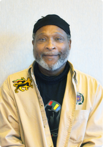 Smiling man in a beige jacket and black cap against a plain light wall