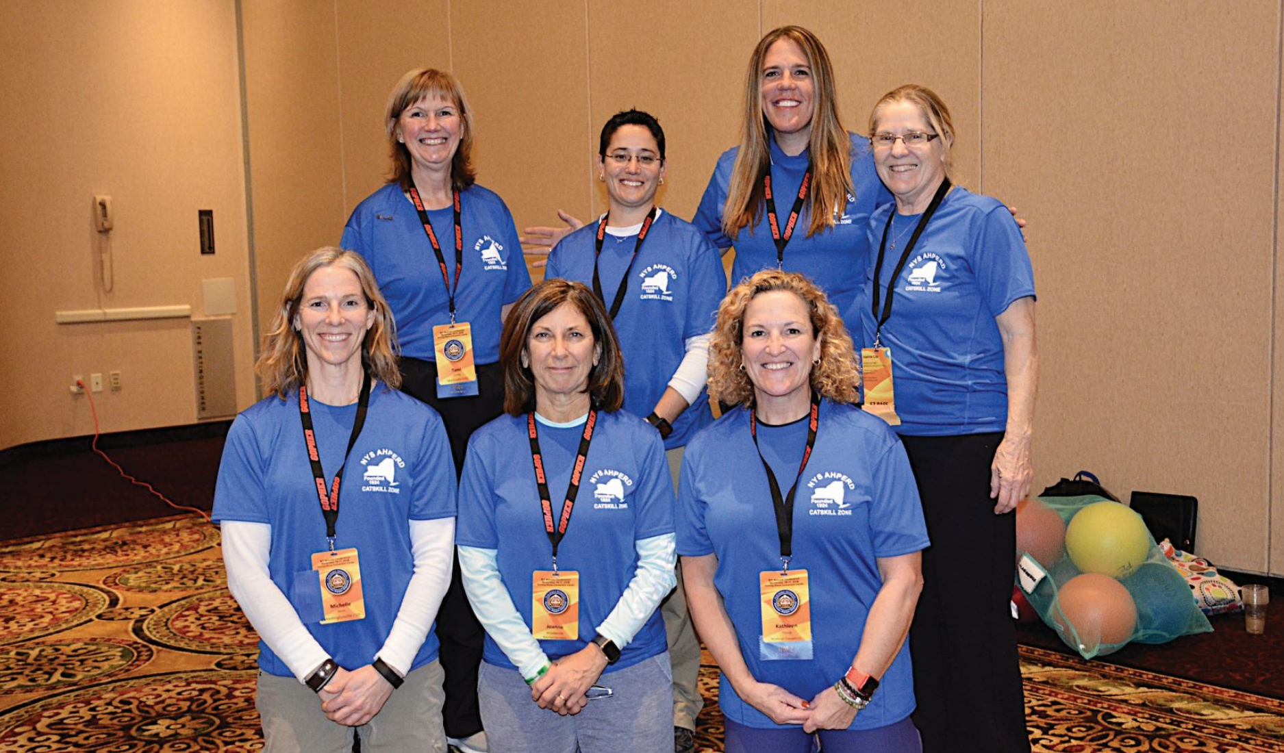 Six women in blue shirts pose with lanyards at a conference in a hotel lobby.