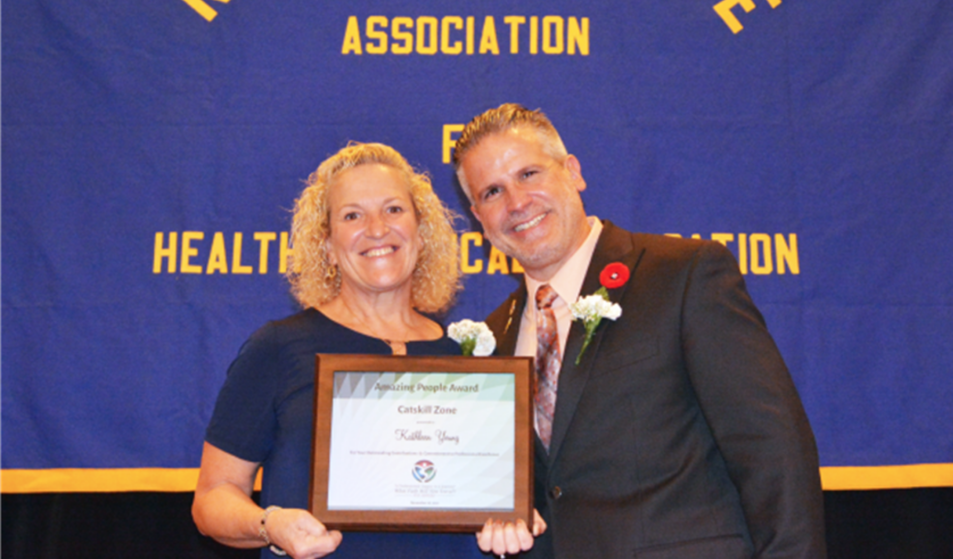 Two people in formalwear hold a framed certificate at an awards event backdrop