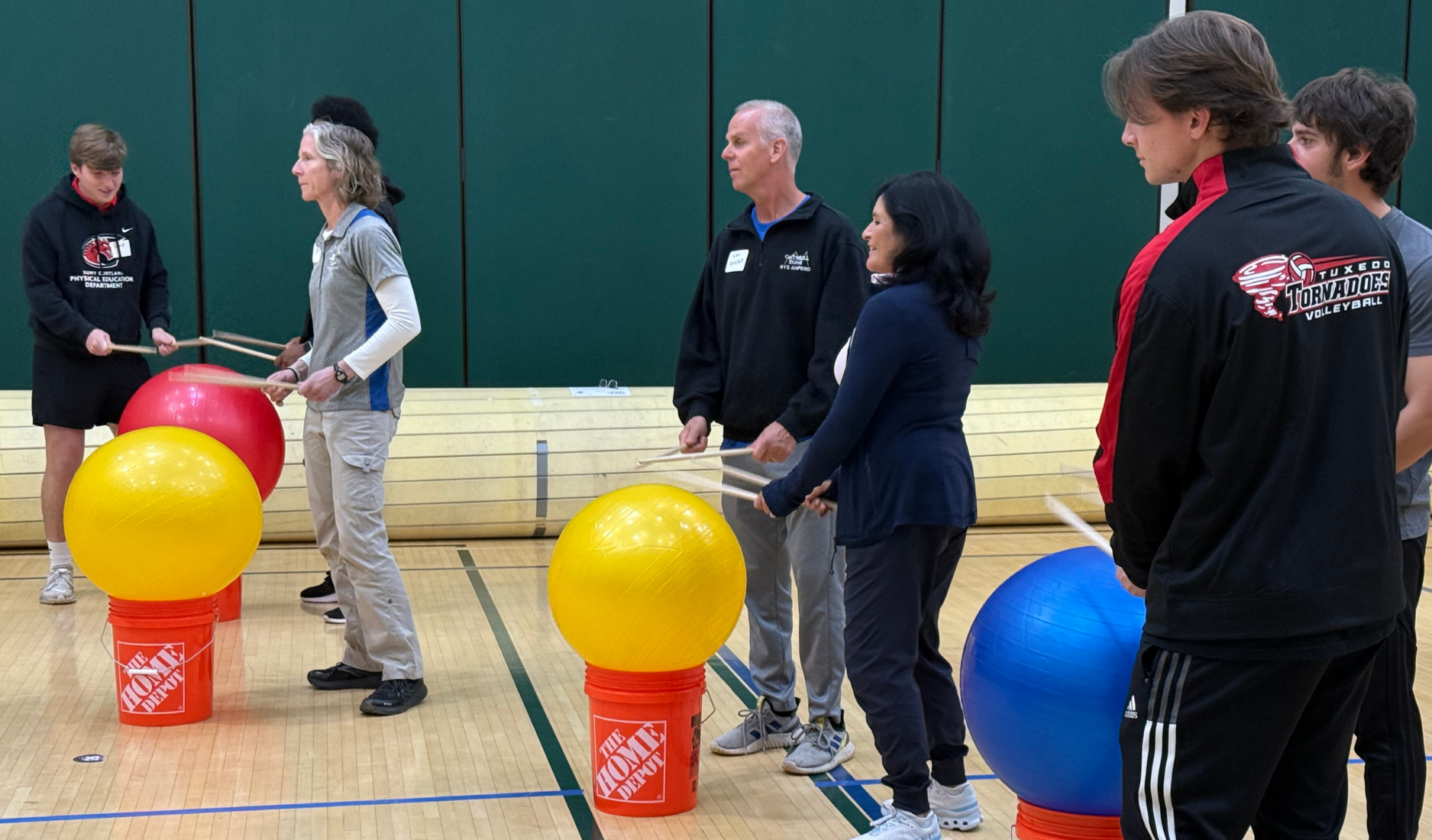 People standing with large red, yellow, and blue balls in a gym, watching an activity.
